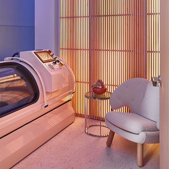 Hyperbaric chamber with orange and pink illuminated wall and cream felt chair on the right of the image