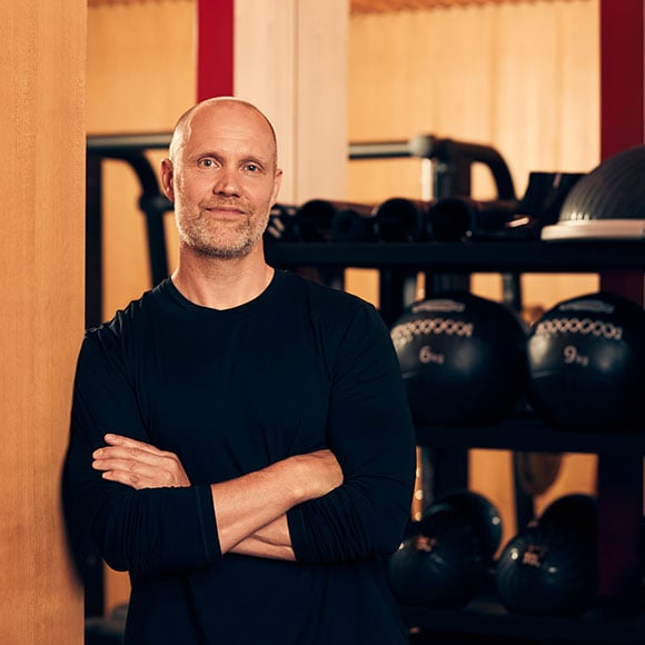 Portrait of Simon Inman wearing a long sleeved black jumper. Gym equipment is located in the background.