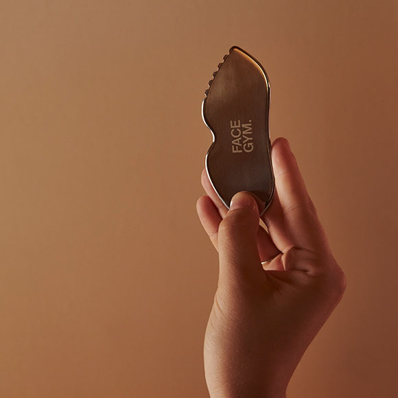 A hand holding up a sleek, metallic Face Gym tool against a neutral, soft brown background. The ergonomic design features smooth, curved edges and the branding 'Face Gym' is visible on the surface of the tool, which is designed for facial massage and sculpting.