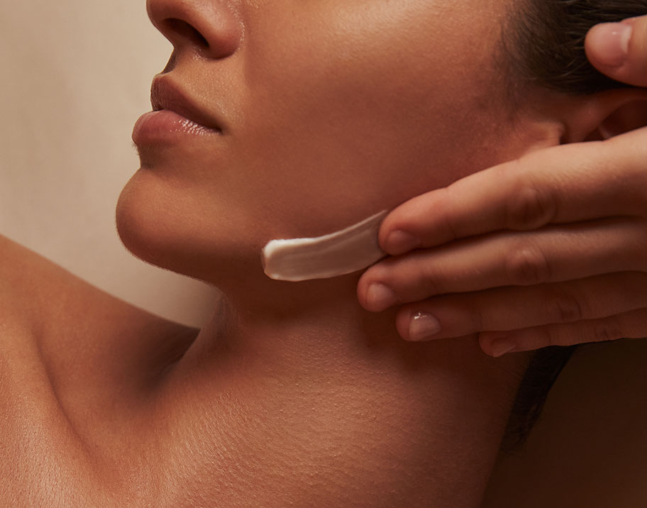 A close up of a hand applying cream to a woman's face. Her head is turned to the side.