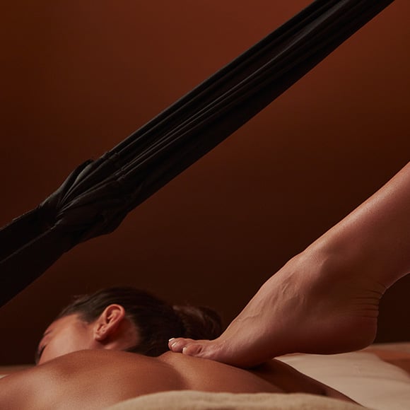 A woman laying down receiving a Ashiatsu massage with a  therapist's foot on her back. A rope stretches out above.