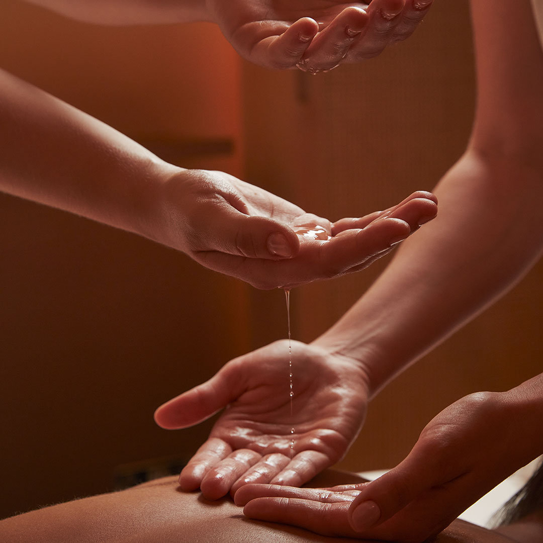 Signature Massage treatment, Four Hands. An image of two practictioners' sets of hands, pouring oil and massaging someone