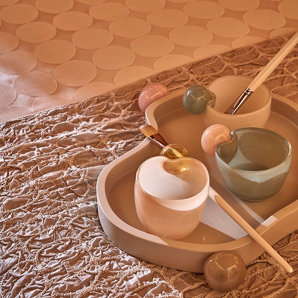 Close-up of a luxurious spa treatment setting featuring delicate pastel-colored ceramic bowls, brushes, and a palette tray. The arrangement sits atop a textured fabric, creating a calming and serene ambiance, perfect for a relaxing experience