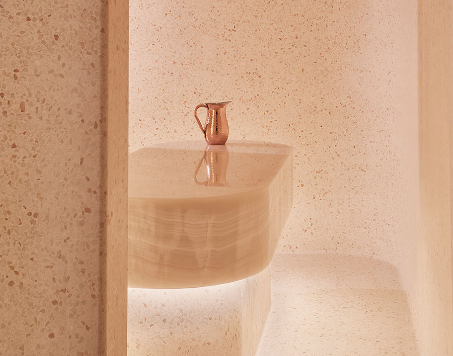 Image of a luxurious Hammam room featuring smooth white onyx surfaces. The room has soft, ambient lighting that highlights the elegant curves of a central onyx wash basin, reflecting a copper pitcher resting on top. The walls are finished with a delicate, speckled texture, enhancing the tranquil and refined atmosphere